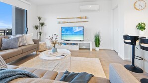 Una sala de estar moderna con sofá, mesa de centro y televisor. Hay plantas, un reloj y una zona de bar.