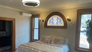 Un dormitorio con una cama grande, un vitral sobre la cama, una puerta de madera y una ventana con vistas al exterior.