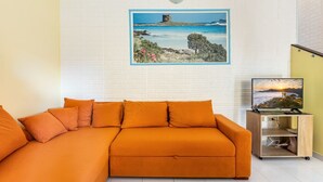 A room with a sectional sofa, a painting of a coastal landscape, a television on a wooden stand, and a white brick wall. - GuestHost - Bright 62sqm apartment, ideal for 3 people, located on the ground floor of a building WITHOUT a lift (two flights of stairs to access it).The accommodation is located in the La Collina residence, in Villasimius, in about 13 minutes by car y (Villasimius)