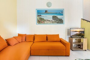 Une pièce comprenant un canapé d’angle, un tableau représentant un paysage côtier, un téléviseur posé sur un meuble en bois et un mur en briques blanches.