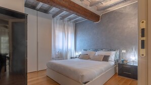 Un dormitorio con una cama grande, piso de madera, techo con vigas a la vista, una mesita de noche y una ventana con cortinas.