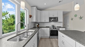Une cuisine moderne avec des meubles blancs, un évier en acier inoxydable, un four à micro-ondes et un four traditionnel. Deux luminaires suspendus et une fenêtre offrent une vue sur la verdure à l’extérieur.