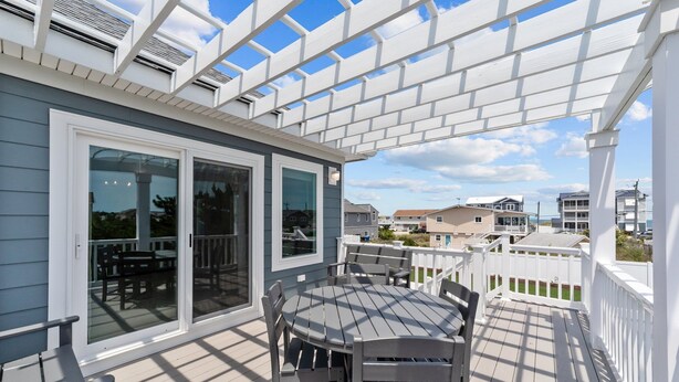 Une terrasse extérieure couverte, dotée d’une table et de chaises, d’une pergola au-dessus et offrant une vue sur les maisons voisines.