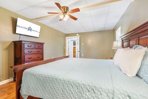 A bedroom with a bed, a wooden dresser, a ceiling fan, and a TV mounted on the wall.