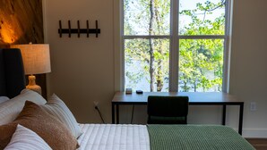 A bedroom with a bed, bedside table, lamp, and a window with a view of trees. - Lakeside Cottage in the Treetops - 2 bedrooms with amazing view (Jacksons' Gap)