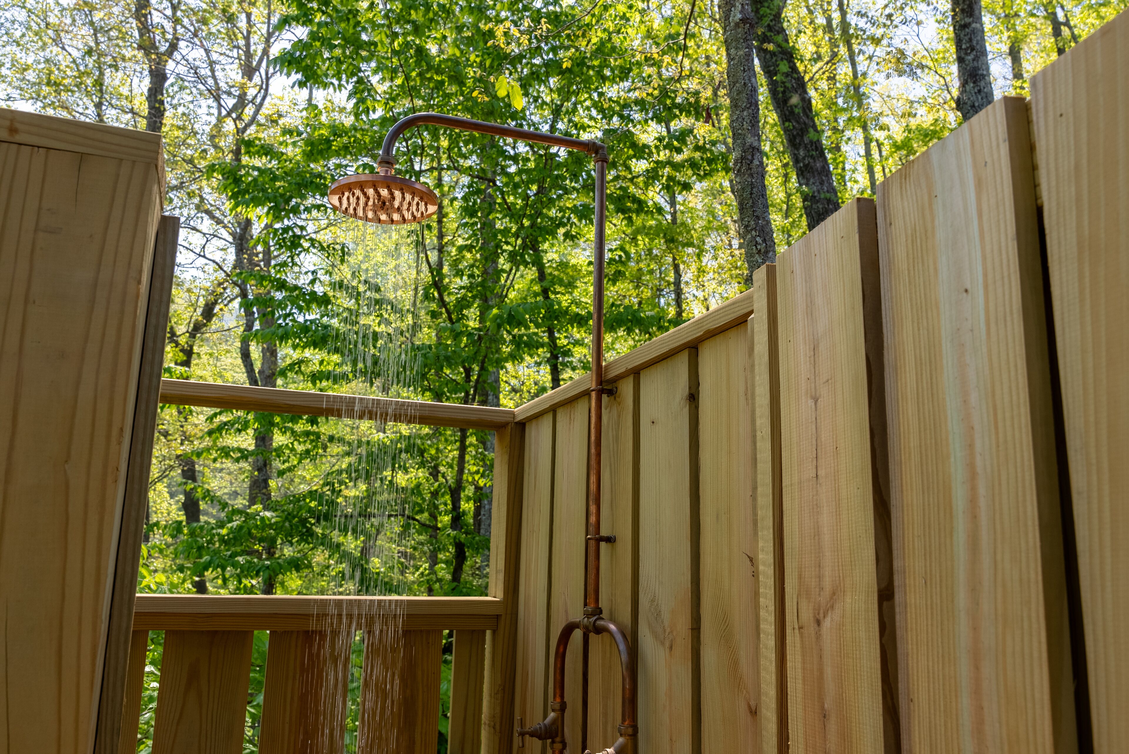 Une douche extérieure en bois, dotée d’un pommeau de douche à effet pluie, entourée d’une clôture en bois et d’arbres.