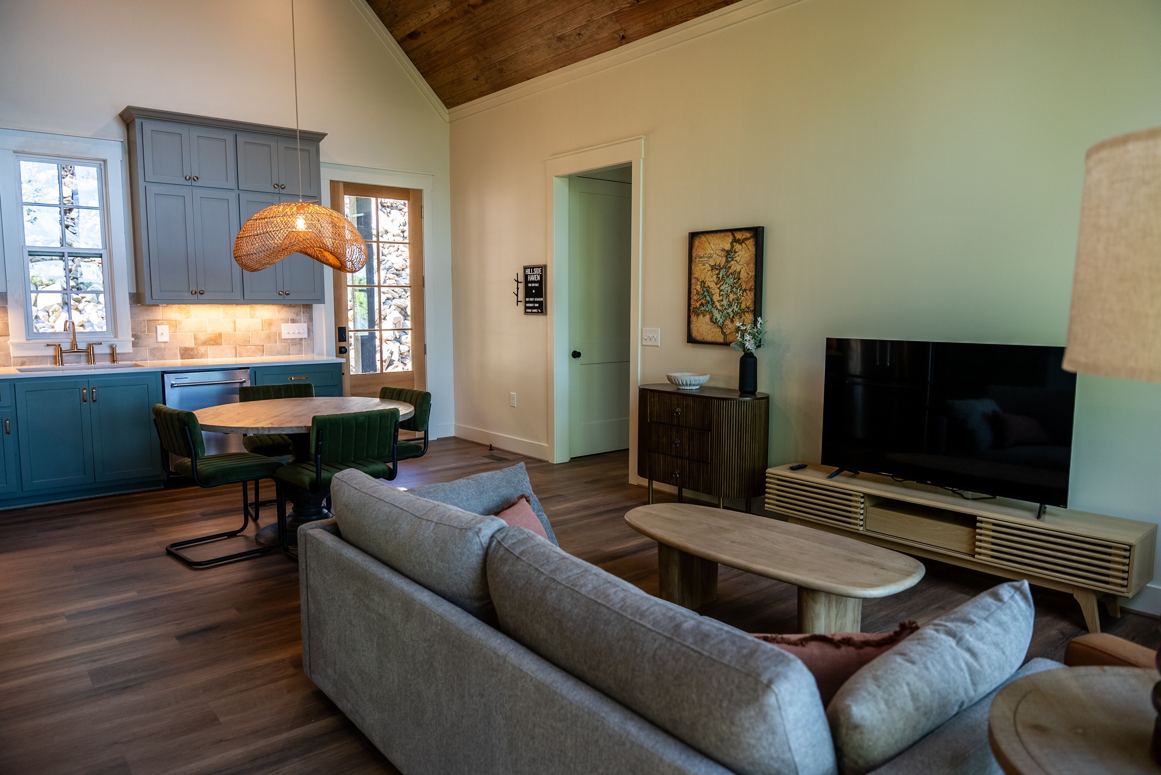 A modern living room with a sofa, coffee table, and TV. There is a dining area with chairs and a table. The kitchen has blue cabinets and a hanging light fixture.