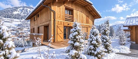 A wooden chalet with a wooden deck, surrounded by snow-covered trees and mountains.