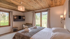 A bedroom with a wooden ceiling, a bed with white linens, a desk, and a TV mounted on the wall.