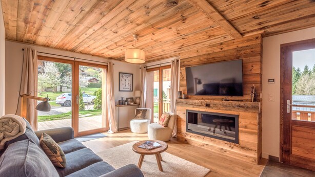 A cozy living room with a wooden ceiling, a large flat-screen TV mounted on a wooden wall, a fireplace, and a sofa with cushions.