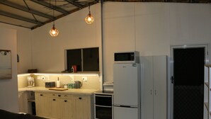Eine kleine Küche mit weißen Schränken, einer Mikrowelle, einem Kühlschrank und einer Kaffeemaschine. Über der Arbeitsplatte hängen Pendelleuchten.