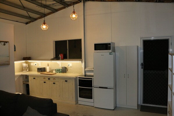 A compact kitchen with white cabinets, a microwave, a refrigerator, and a coffee maker. There are pendant lights above the counter. - Beach Shed Toogoom (Toogoom)