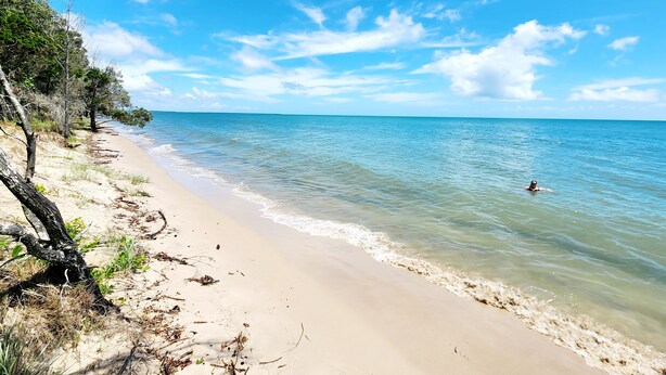 Ein Strand mit klarem, blauem Wasser, einem Sandufer und einer schwimmenden Person.