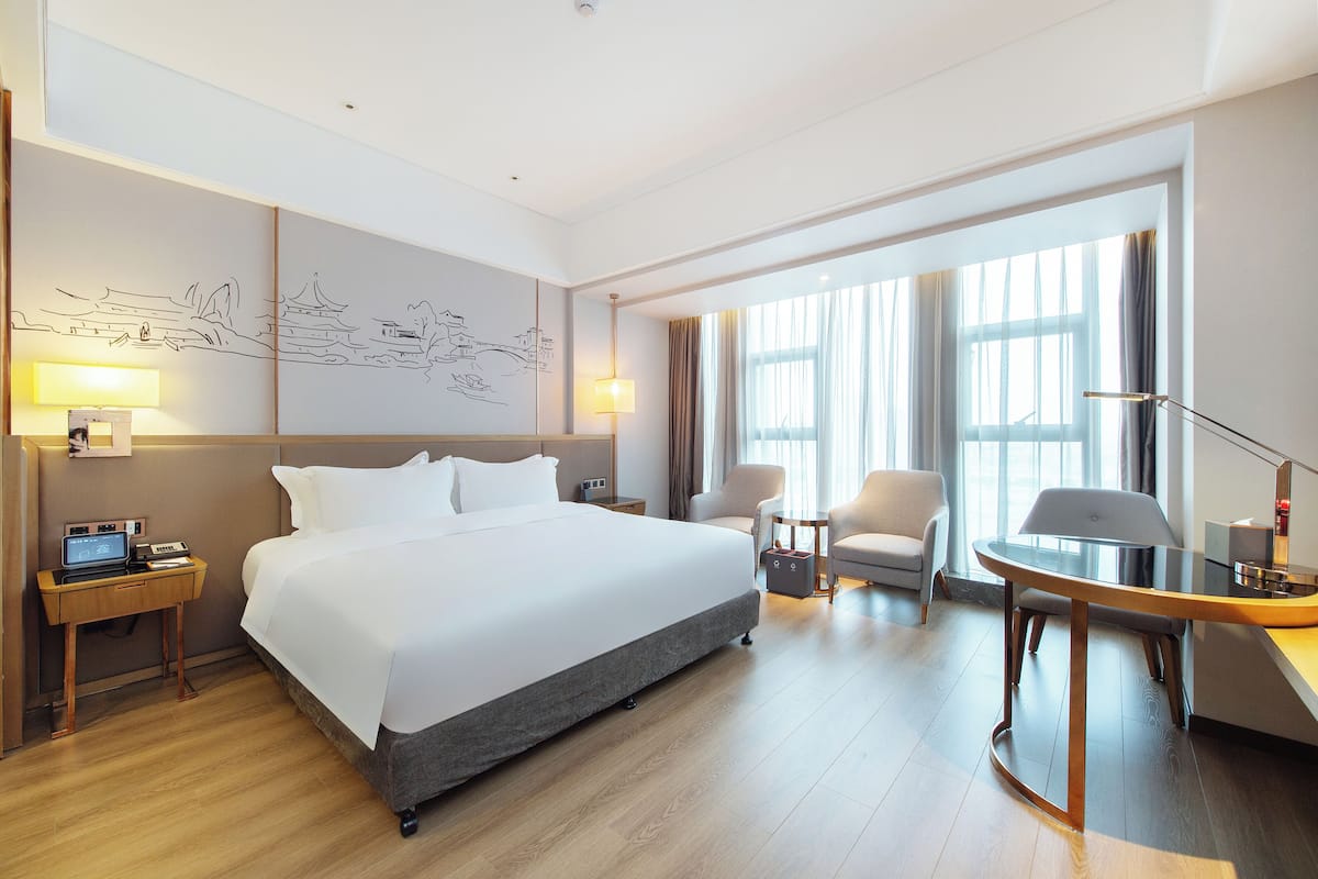 A modern hotel room with a large bed, a desk with a chair, a small table, and a large window with curtains.