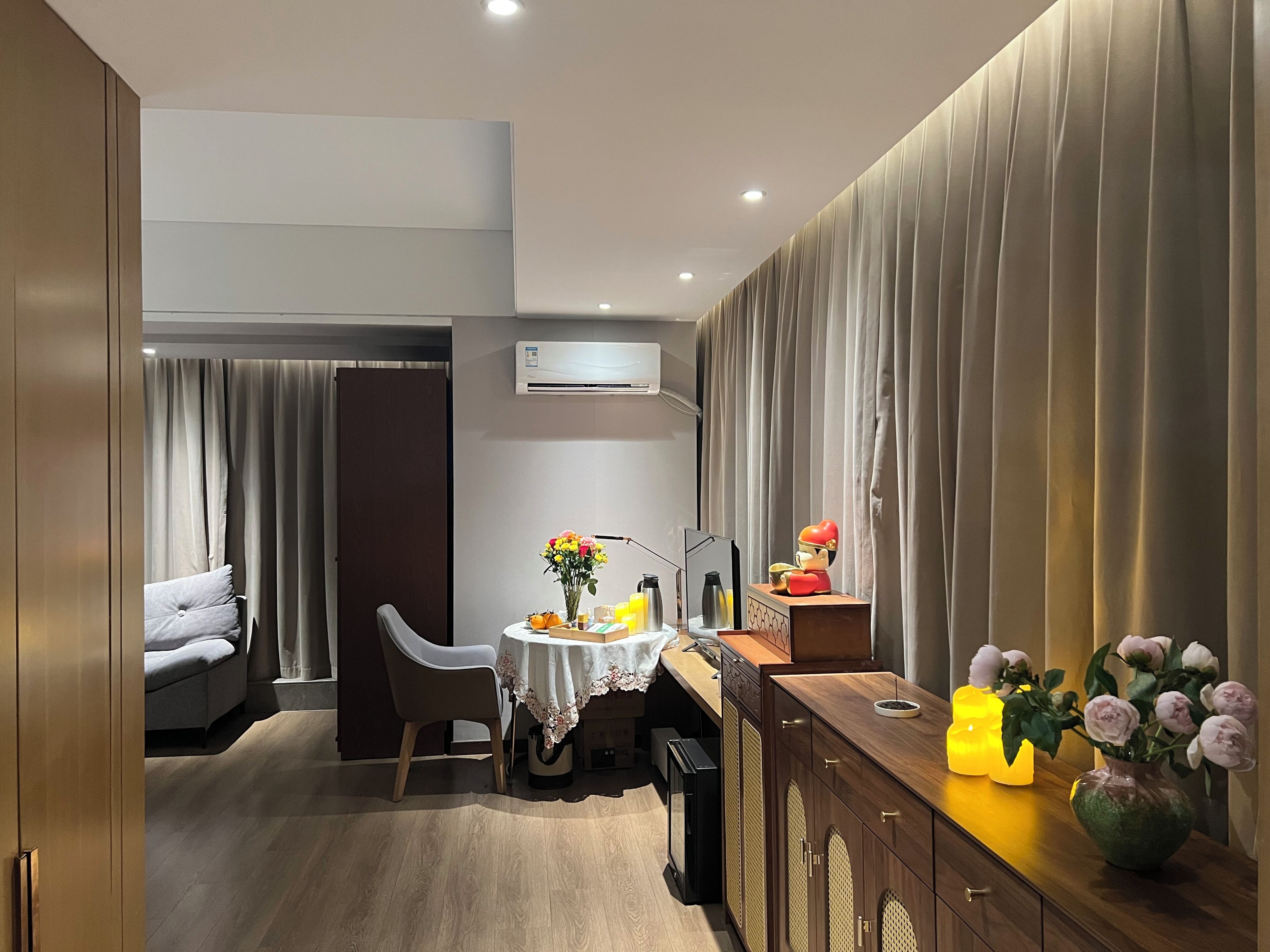 A modern hotel room with a dining area, a sofa, a table with a vase of flowers, and a cabinet with decorative items.