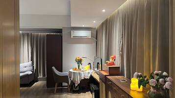 A modern hotel room with a dining area, a sofa, a table with a vase of flowers, and a cabinet with decorative items.