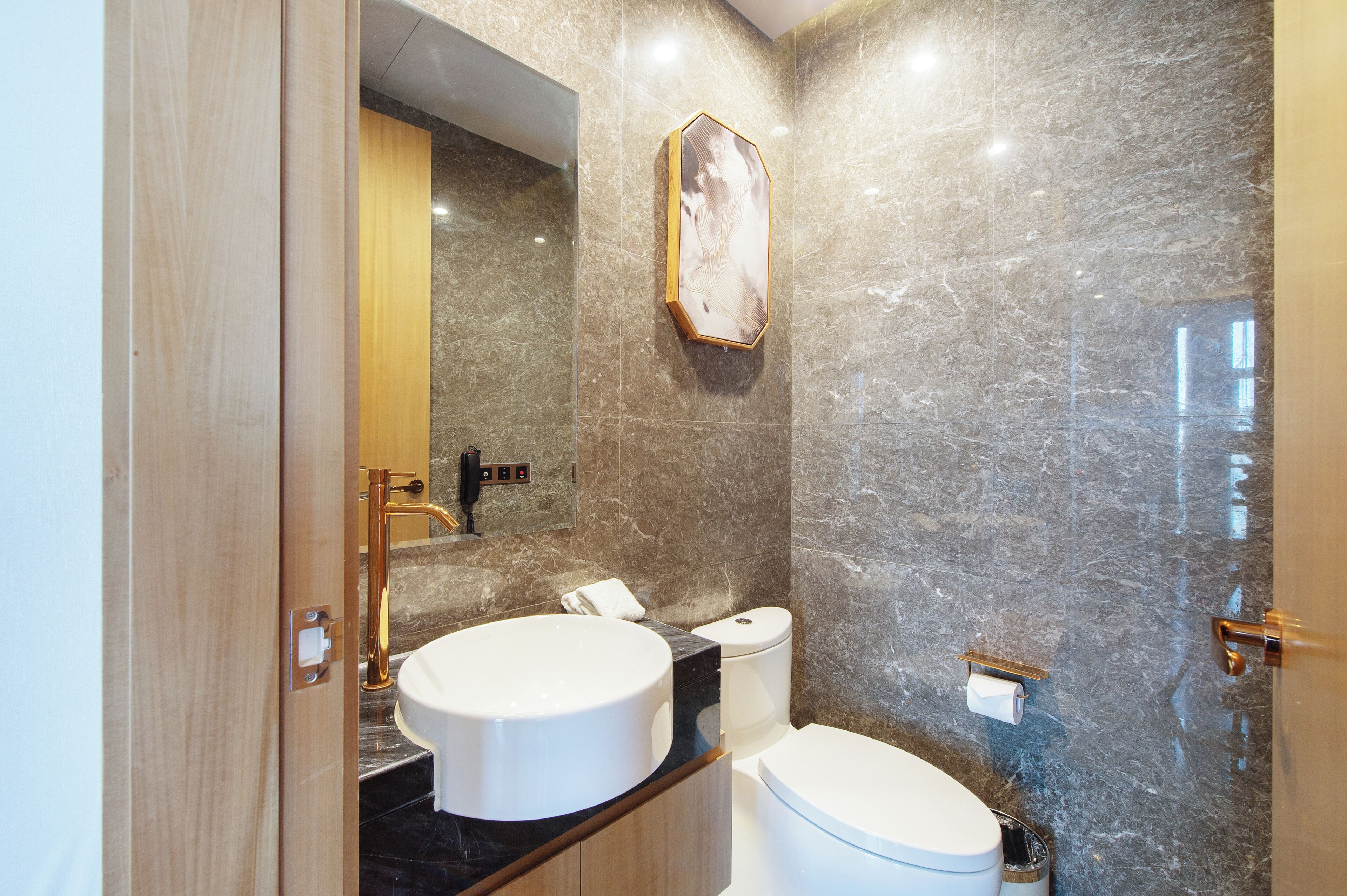 A modern bathroom with a marble wall, a wooden vanity, a round sink, and a toilet.