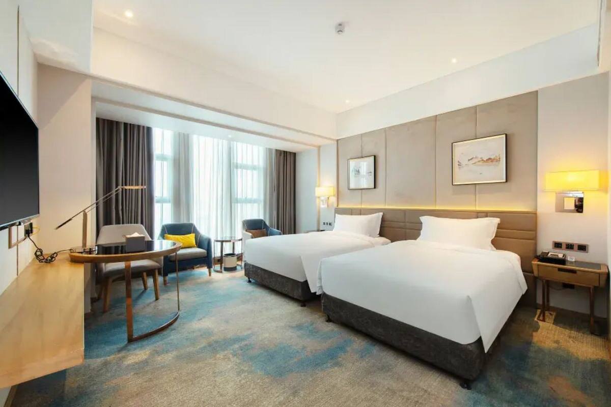 A hotel room with two beds, a desk, a chair, a TV, and a large window with curtains.