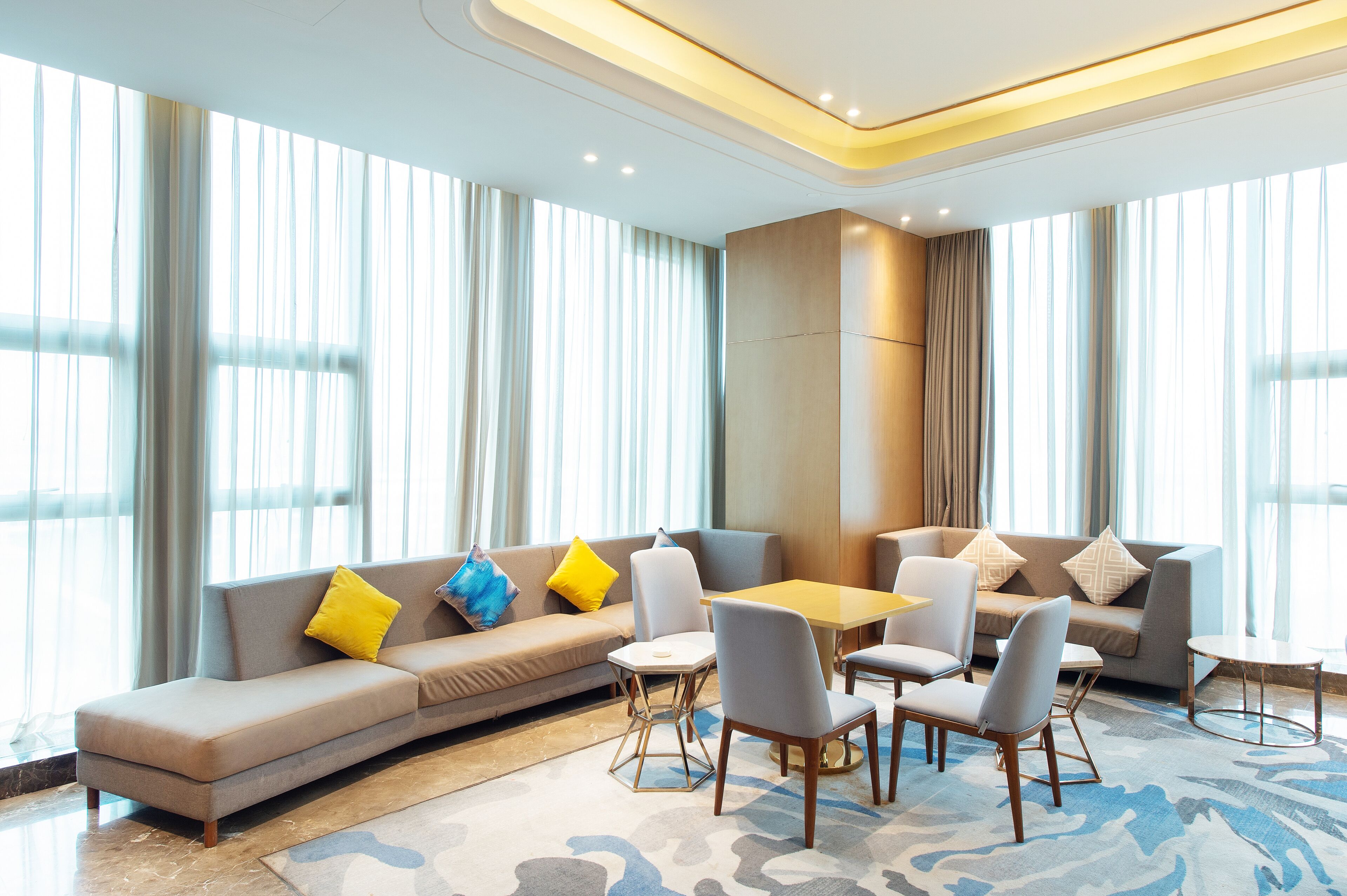 A modern hotel room with a large window, a sofa set, a dining table, and a small side table.