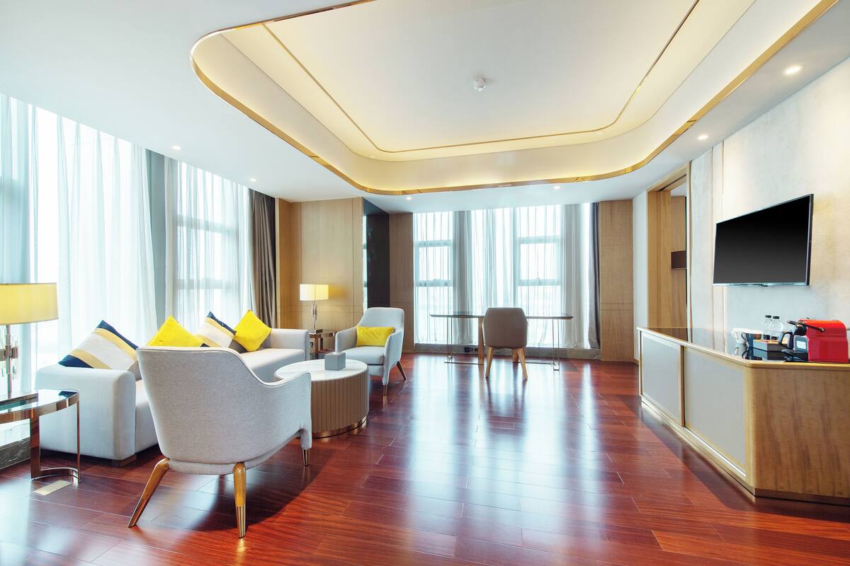A modern hotel lobby with a wooden floor, a seating area with sofas and armchairs, a television on the wall, and a reception desk.