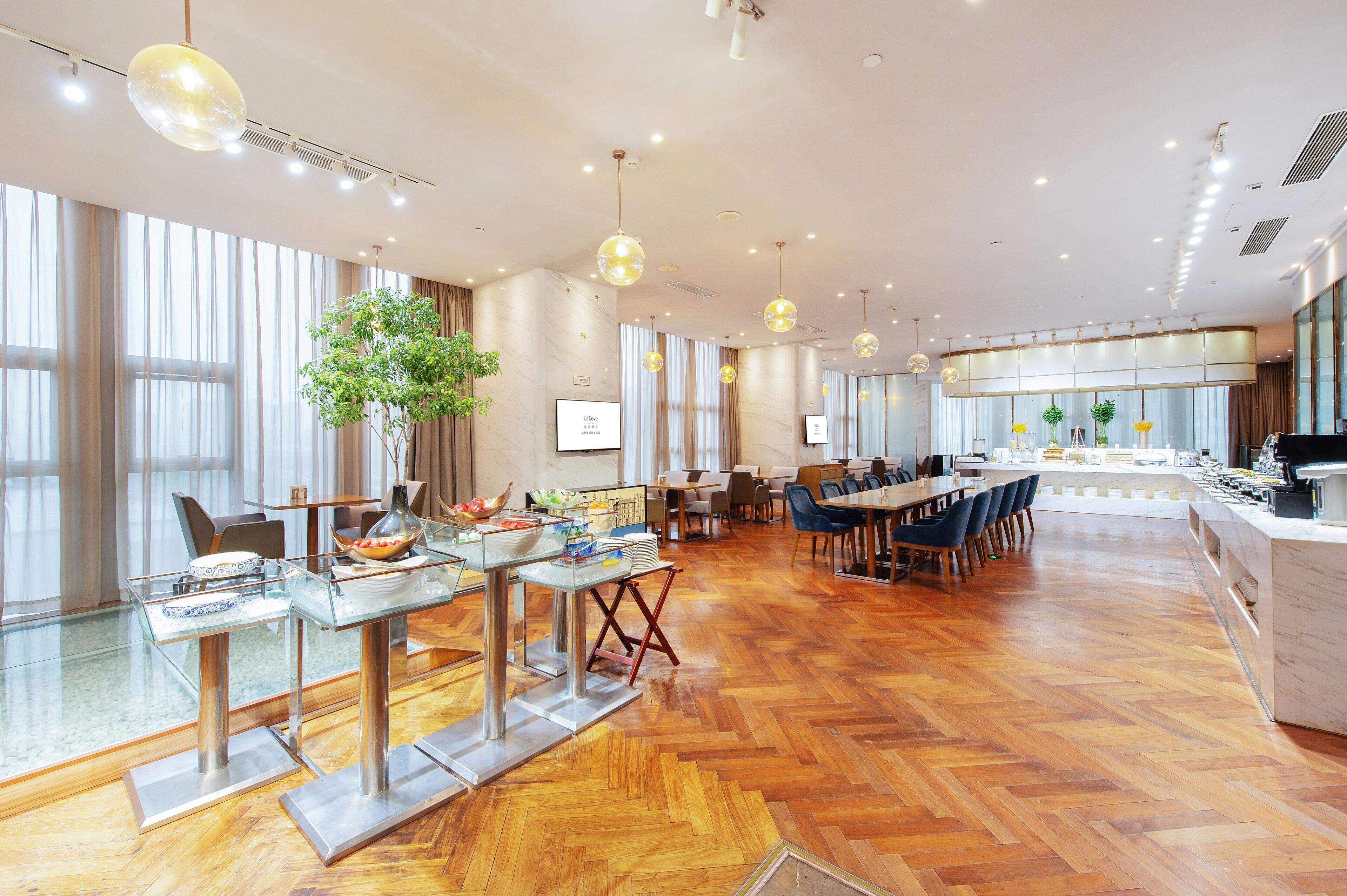 A spacious hotel dining area with modern furniture, large windows, and a central plant.