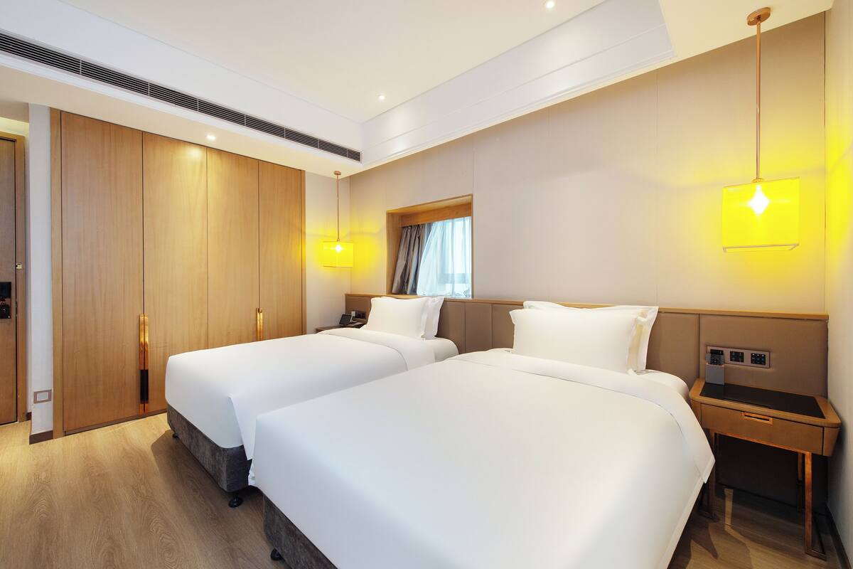 A hotel room with two beds, a wardrobe, a bedside table, and a hanging light fixture.