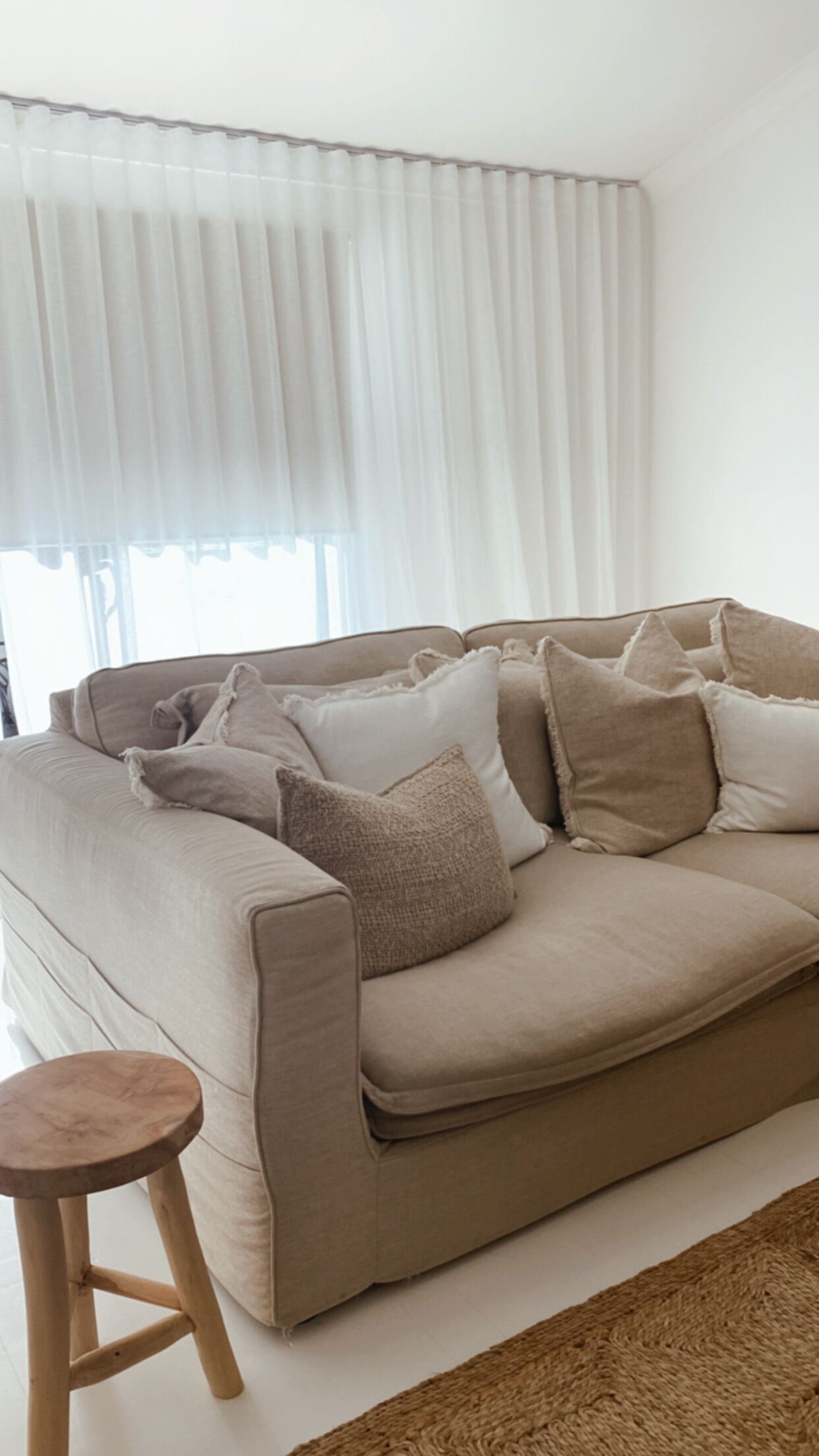 Ein beiges Sofa mit vielen Kissen, ein Holzschemel und ein weißer Vorhang.