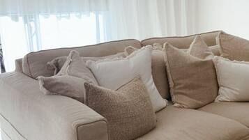 Ein beiges Sofa mit vielen Kissen, ein Holzschemel und ein weißer Vorhang.