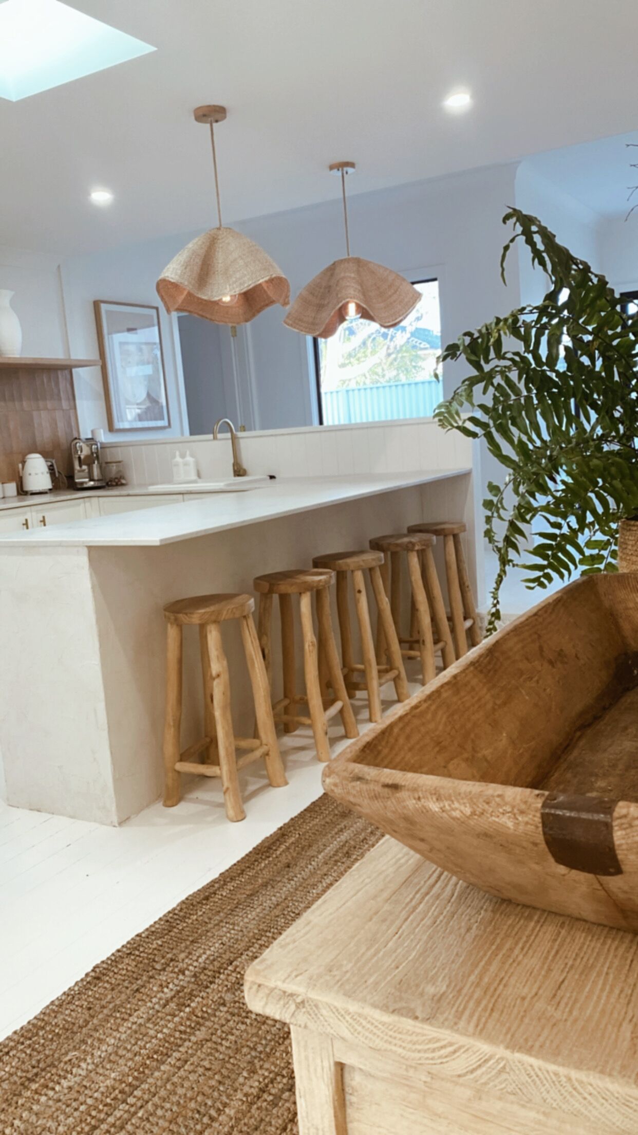 A modern kitchen with a central island, wooden stools, pendant lighting, and a large plant.