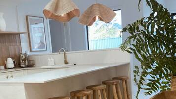 A modern kitchen with a central island, wooden stools, pendant lighting, and a large plant.