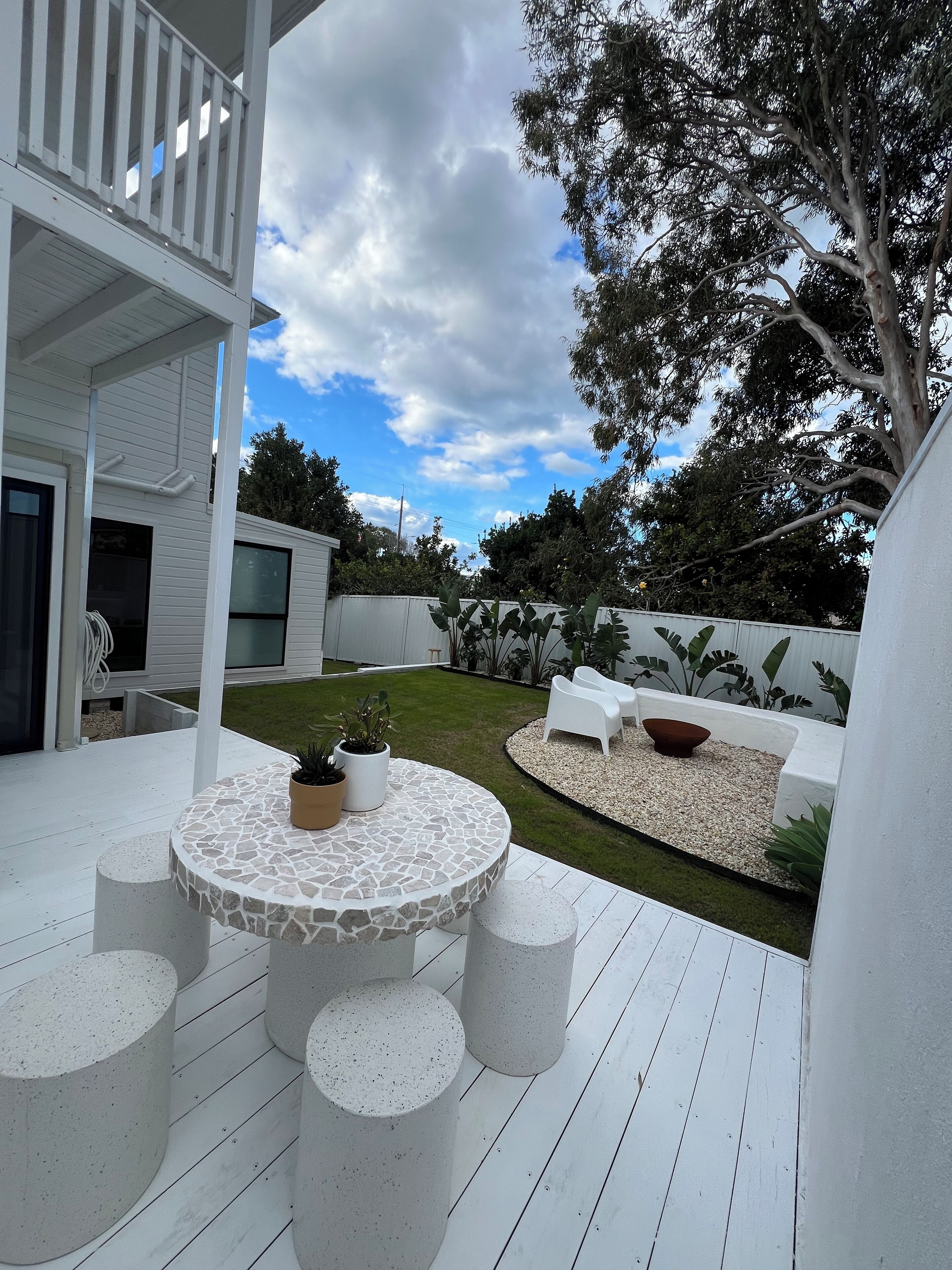 A modern outdoor patio with a round stone table surrounded by white stools, a white fence, and a well-maintained lawn.