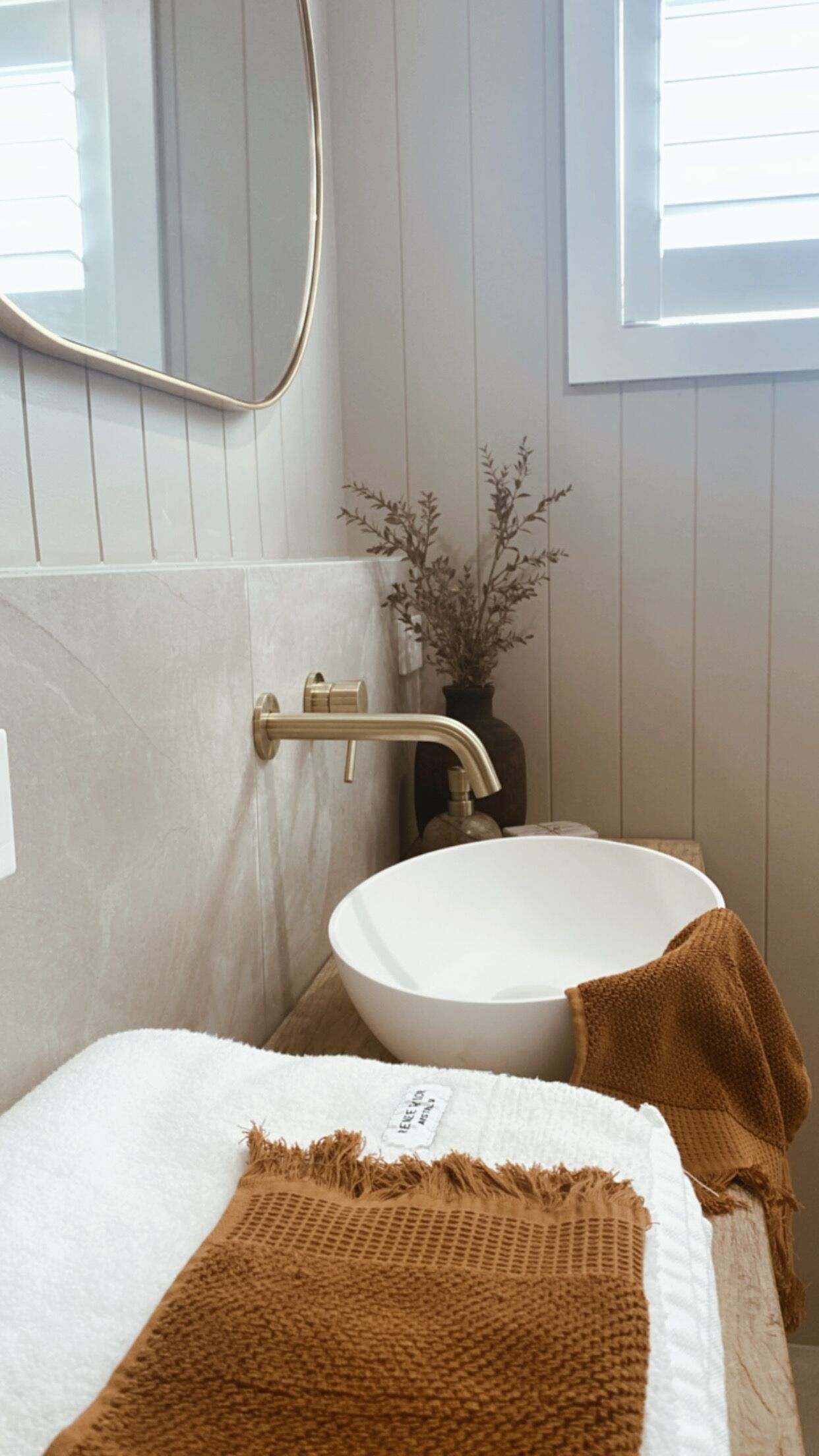 A bathroom with a white sink, a gold faucet, a towel, and a vase with dried plants.