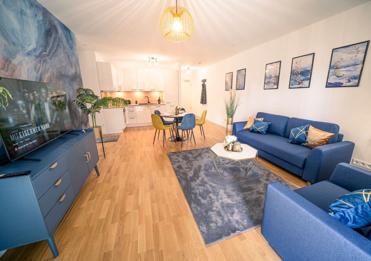 Ein modernes Wohnzimmer mit einer blauen Sofagarnitur, einem Essbereich mit gelben Stühlen und einer Küche im Hintergrund.