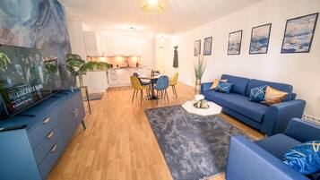 Ein modernes Wohnzimmer mit einer blauen Sofagarnitur, einem Essbereich mit gelben Stühlen und einer Küche im Hintergrund.
