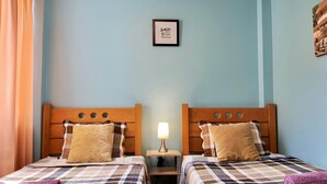 Habitación doble con dos camas, cada una con cabecero de madera, colcha acolchada y toalla morada. Una mesita de noche con lámpara se encuentra entre las camas.