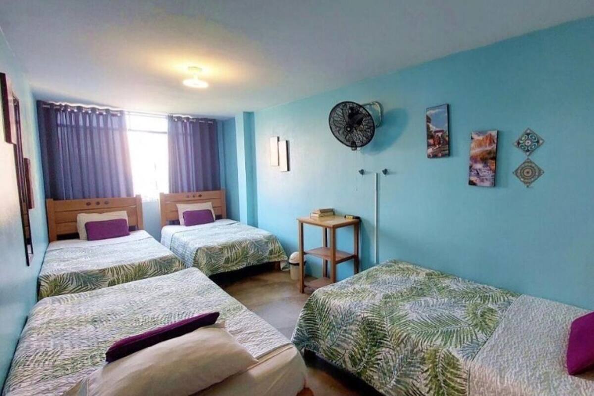 A room with two single beds, a small table, a standing fan, and a window with curtains.