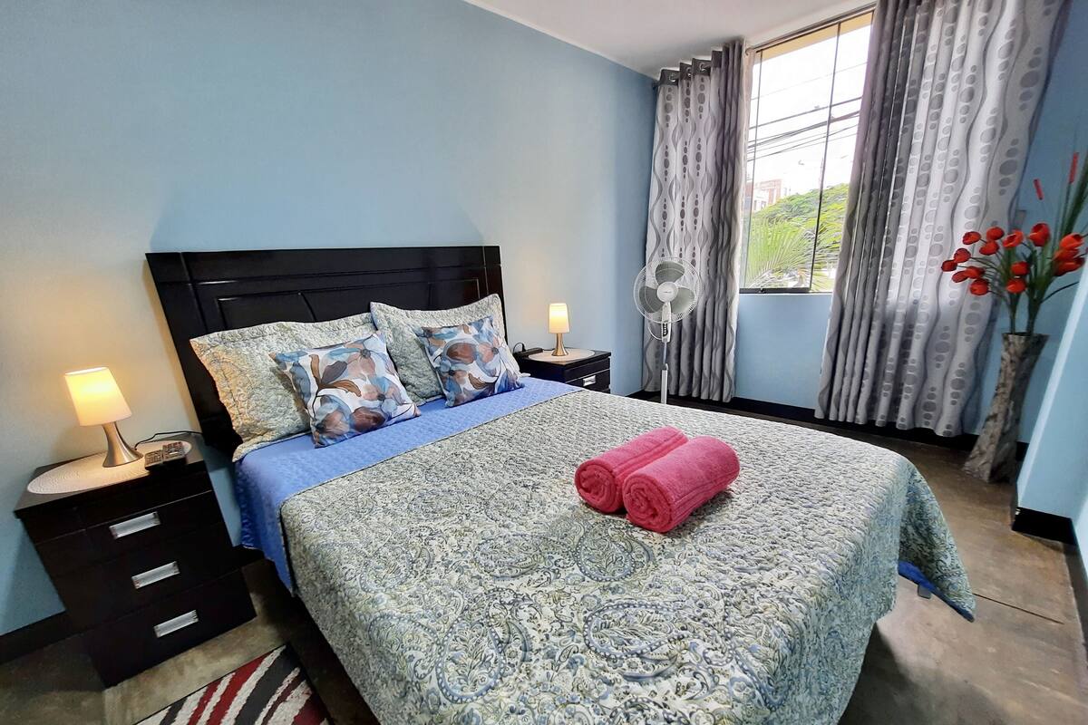 A neatly made bed with a patterned bedspread, two pillows, and a rolled towel. There is a bedside table with a lamp and a fan. The room has light blue walls and a window with curtains.
