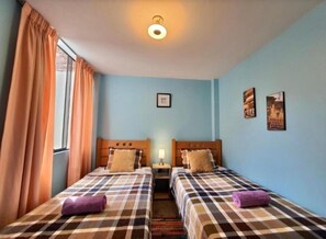 A room with two beds, each with a checkered bedspread and a pillow, a nightstand with a lamp, and a window with curtains.