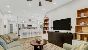 Un salon moderne avec un canapé, une table basse et un téléviseur. On aperçoit une cuisine à l’arrière-plan, avec des meubles blancs, ainsi qu’un coin repas avec des chaises blanches.