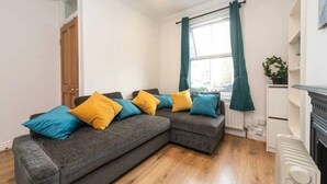 A modern living room with a grey sectional sofa, wooden flooring, a white radiator, and a window with teal curtains. - Charming Period Home near Surbiton with Garden (Greater London)
