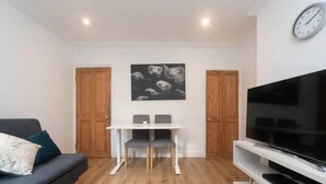 A modern living room with a sofa, a dining table with chairs, a flat-screen TV, and wooden flooring. - Charming Period Home near Surbiton with Garden (Greater London)