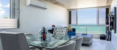 A modern living room with a glass dining table, grey upholstered chairs, a blue sofa, and a view of the ocean through large windows.