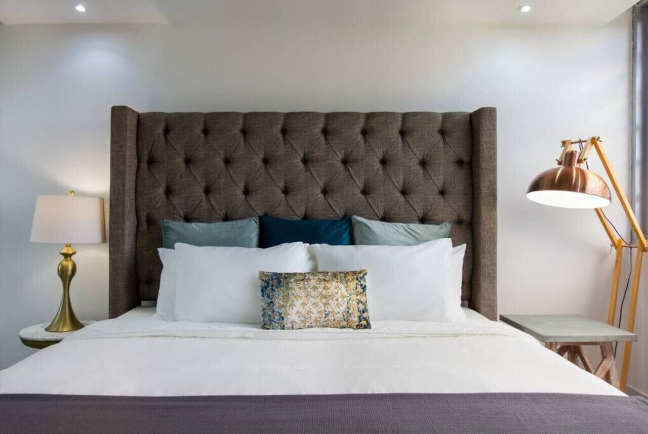 Cama bem arrumada com cabeceira estofada, luminária de cabeceira e uma pequena mesa com abajur.