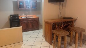 Cocina compacta con estufa, lavamanos, armarios de madera, un televisor montado en la pared y una mesa de madera con taburetes.