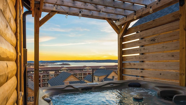 A wooden cabin with a hot tub and a view of a lake and buildings in the background. - Lakeview Cabin w/ Hot Tub, Pool, Decks - Sleeps 20 (Branson)