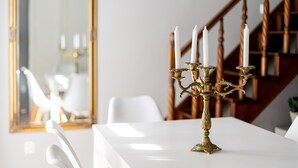 A white table with a brass candelabra holding four white candles, set against a wooden staircase and a mirror reflecting a white chair.