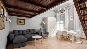 A modern living room with a grey sectional sofa, a round coffee table, and a grey armchair. There is a dining area with a white table and chairs, and a white wardrobe in the background.