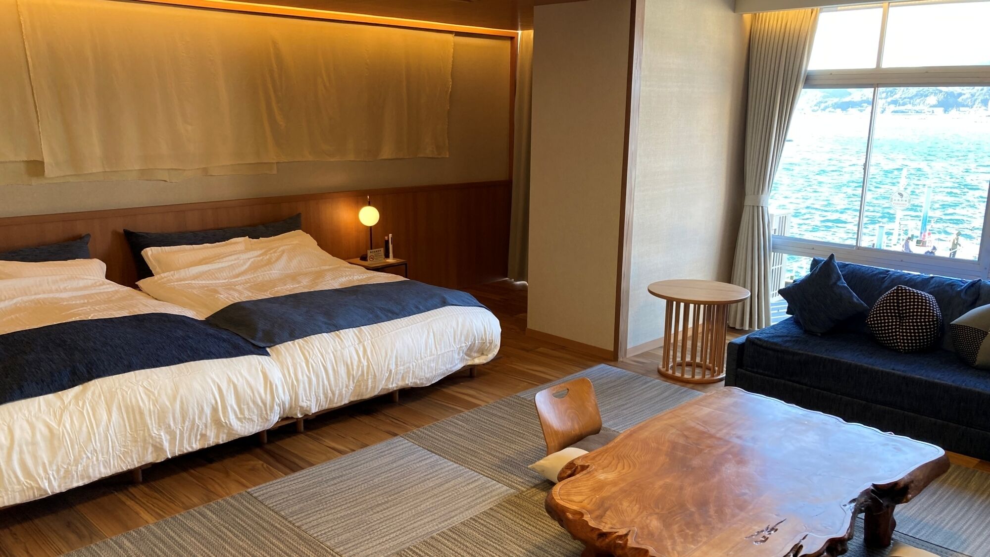 Une chambre d’hôtel avec un lit, un canapé, une table ronde en bois, une table de chevet et une fenêtre donnant sur la mer.