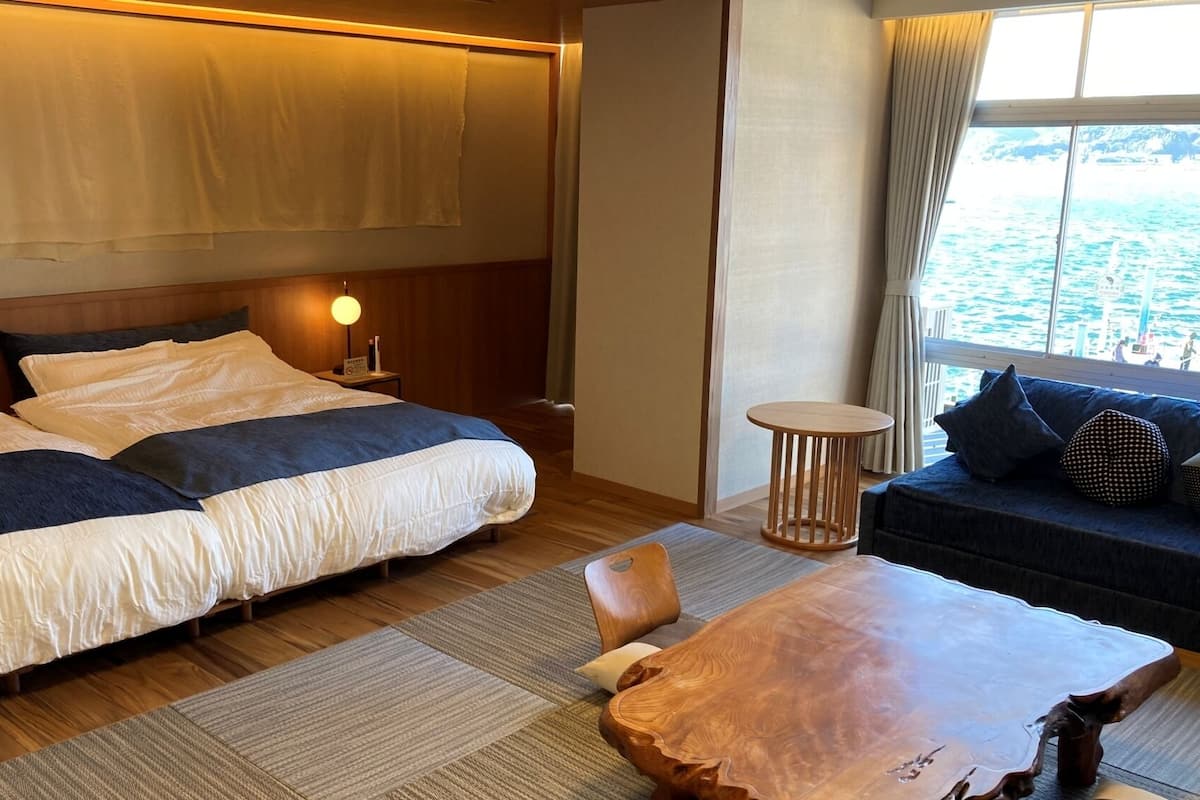 Une chambre d’hôtel avec un lit, un canapé, une table ronde en bois, une table de chevet et une fenêtre donnant sur la mer.