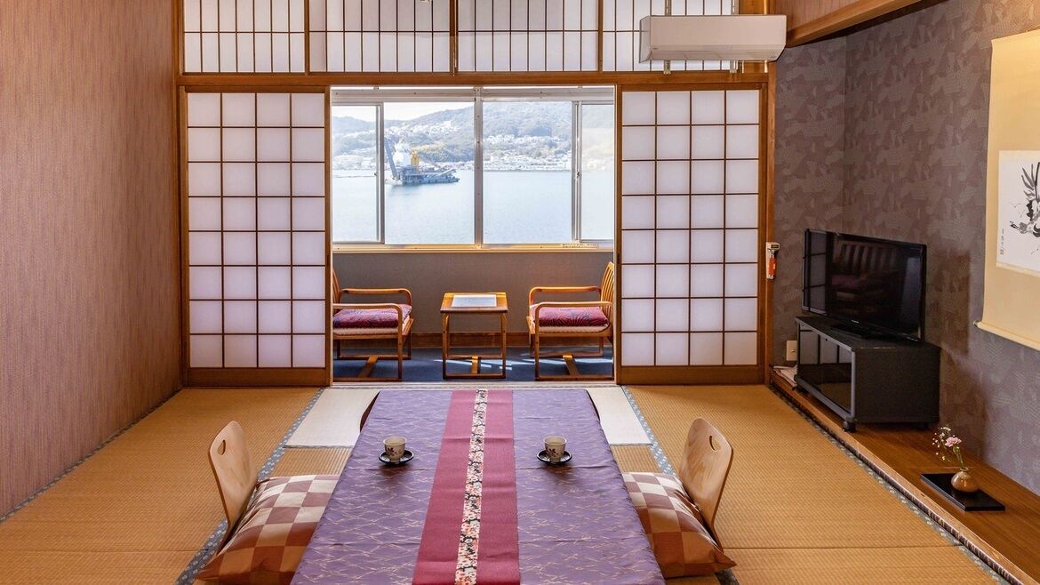 Una habitación japonesa tradicional con suelo de tatami, una mesa baja con cojines, un televisor y vistas a un cuerpo de agua.